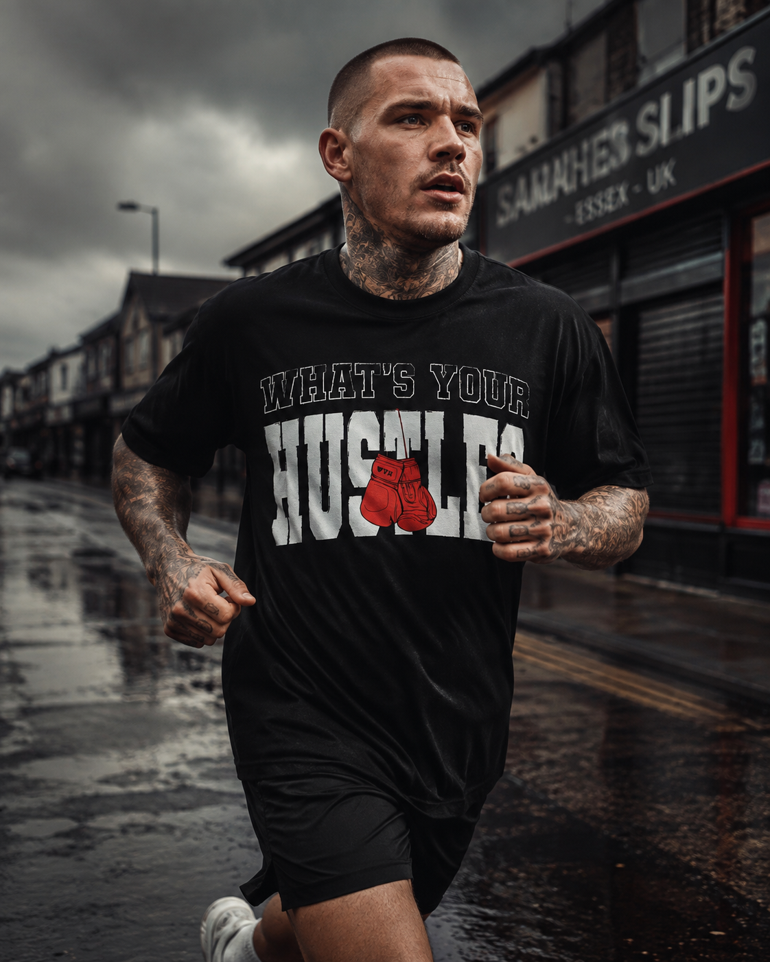 A man running on a wet city street wearing a black graphic t-shirt with the text 'WHAT'S YOUR HUGSLES' and a red boxing gloves graphic, alongside black athletic shorts.