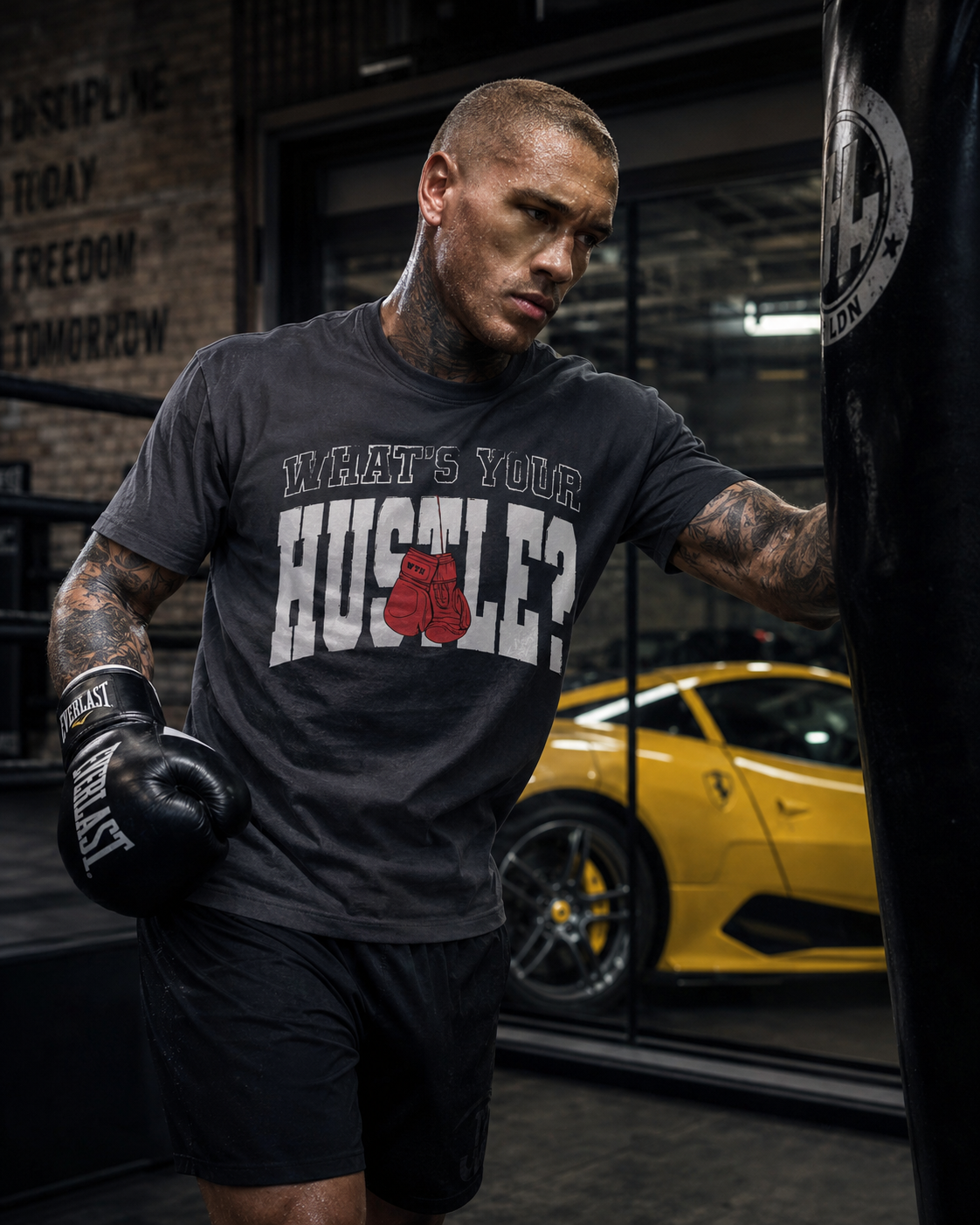 A tattooed man wearing a dark gray t-shirt with a red and white graphic that reads 'WHAT'S YOUR HUSTLE?' and black boxing gloves stands next to a punching bag in a gym, with a yellow sports car visible in the background.