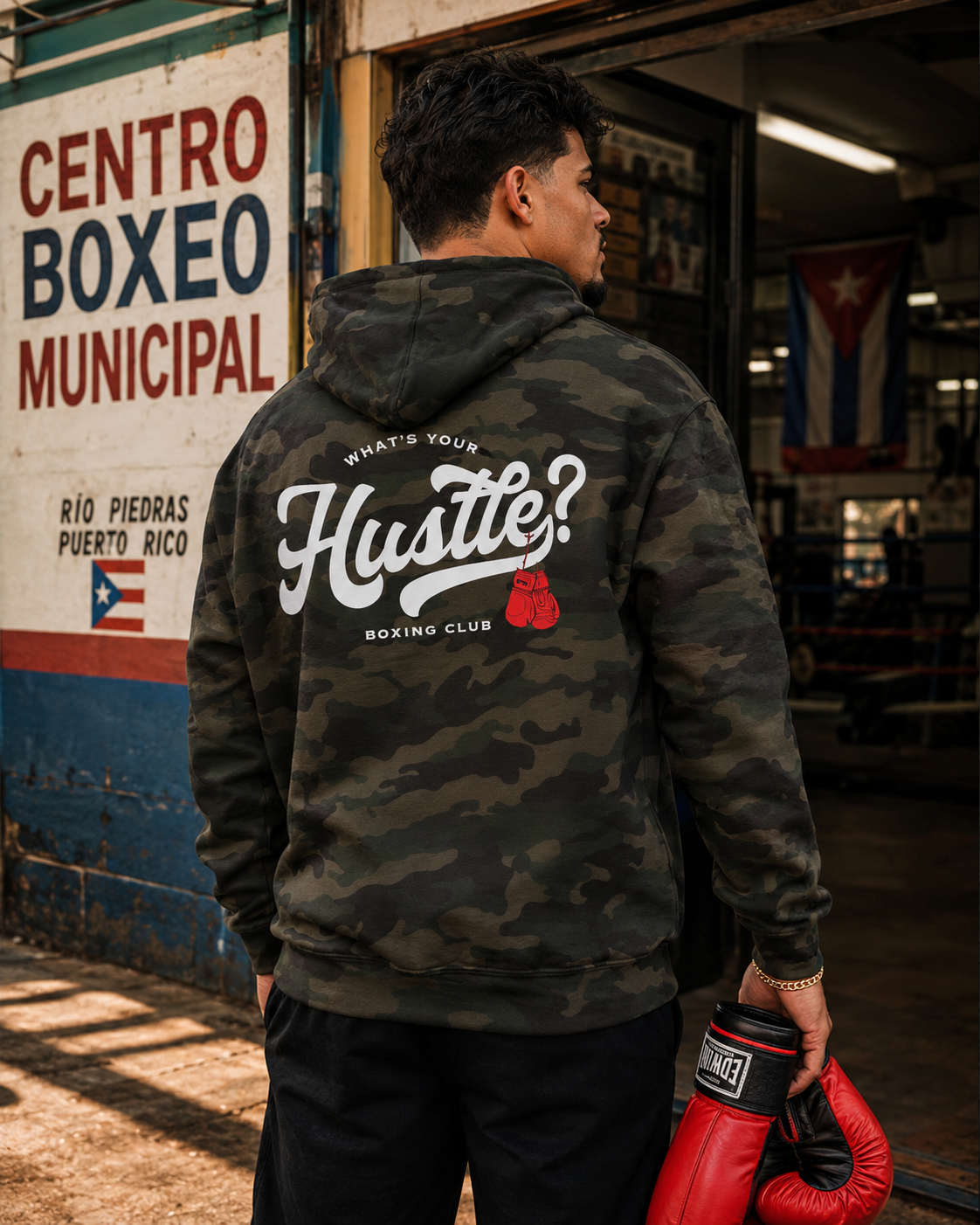 A man in a camouflage hoodie stands in a boxing gym, seen from behind. The hoodie features a large white graphic on the back reading 'Hustle! Boxing Club' with a small red boxing glove accent.