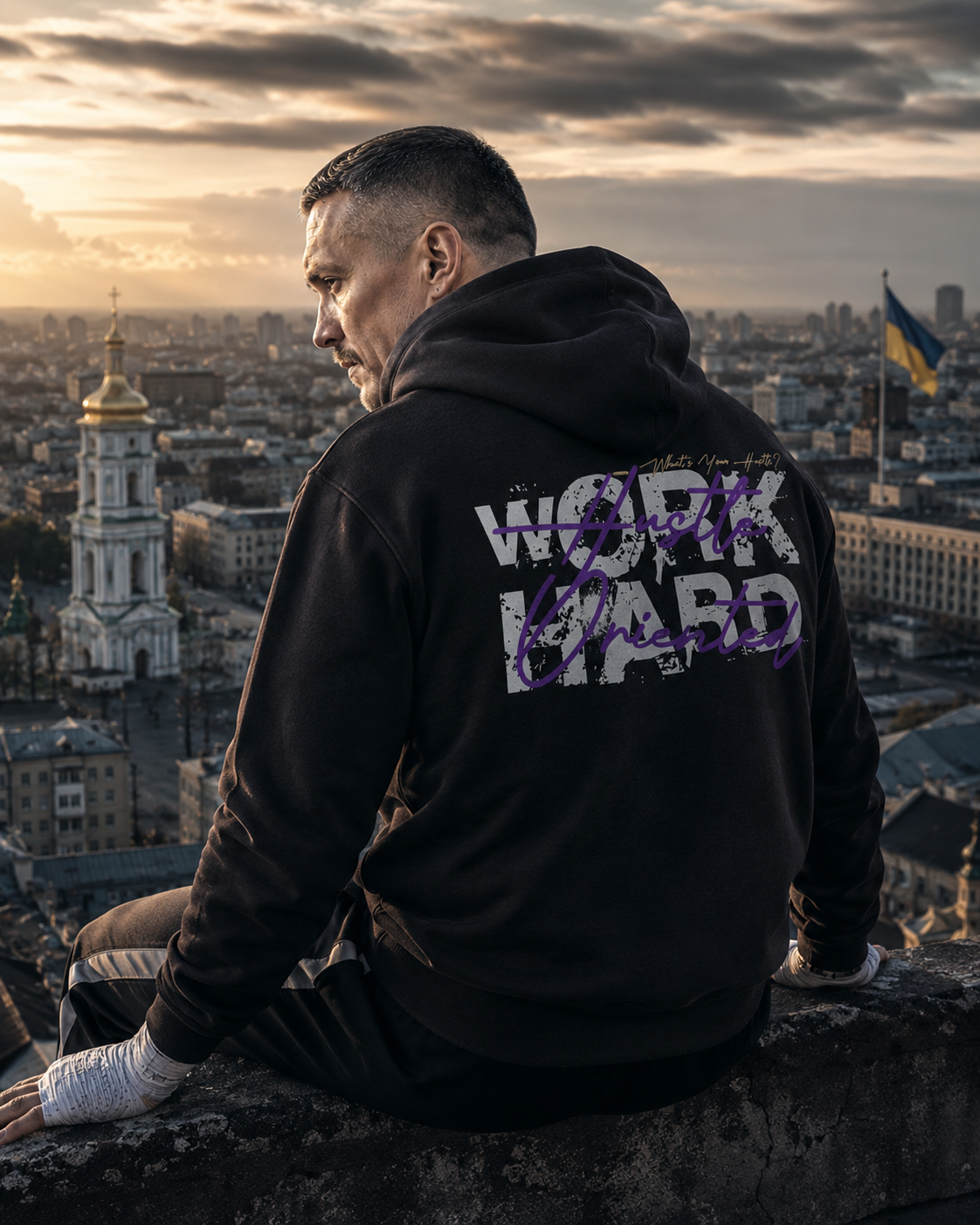 A man wearing a black hoodie with a distressed 'WORK HARD' graphic sits on a stone wall overlooking a city skyline at sunset.