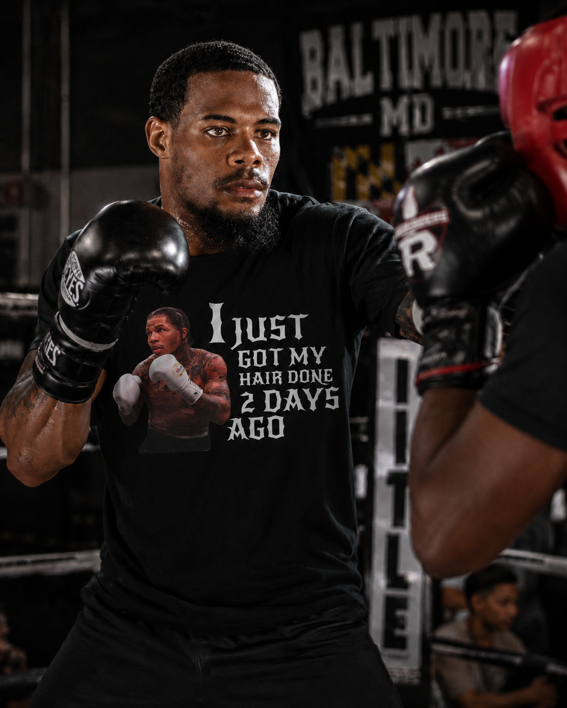 A man in a boxing ring wearing a black graphic t-shirt with the text 'I JUST GOT MY HAIR DONE 2 DAYS AGO' and a photo of a boxer, facing another person wearing red boxing gloves.