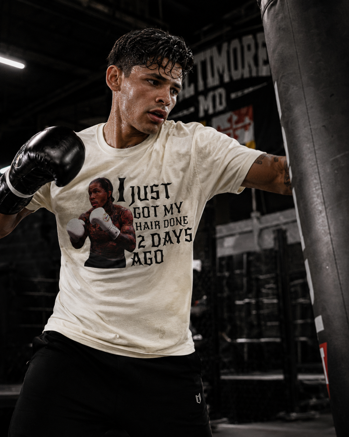 A man in a boxing gym wearing a graphic t-shirt that reads 'I JUST GOT MY HAIR DONE 2 DAYS AGO' with a photo of a boxer, paired with black boxing gloves and punching a heavy bag.