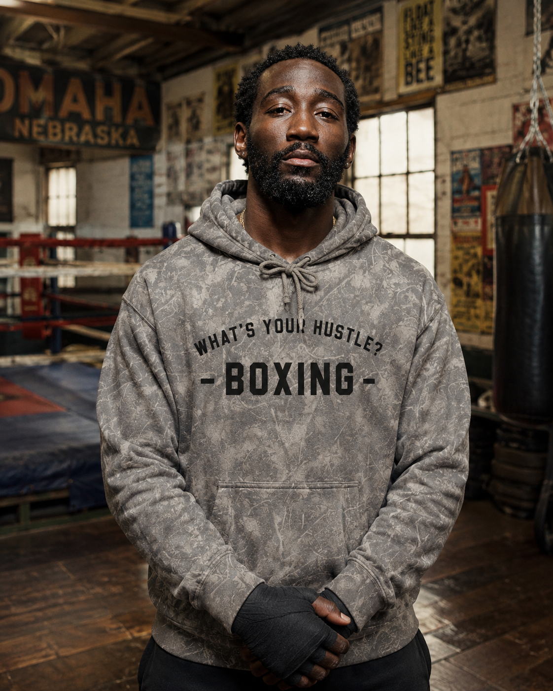 A man wearing a gray camouflage hoodie with 'WHAT'S YOUR HUSTLE - BOXING -' printed on the chest, standing in a boxing gym with a punching bag and boxing rings in the background.