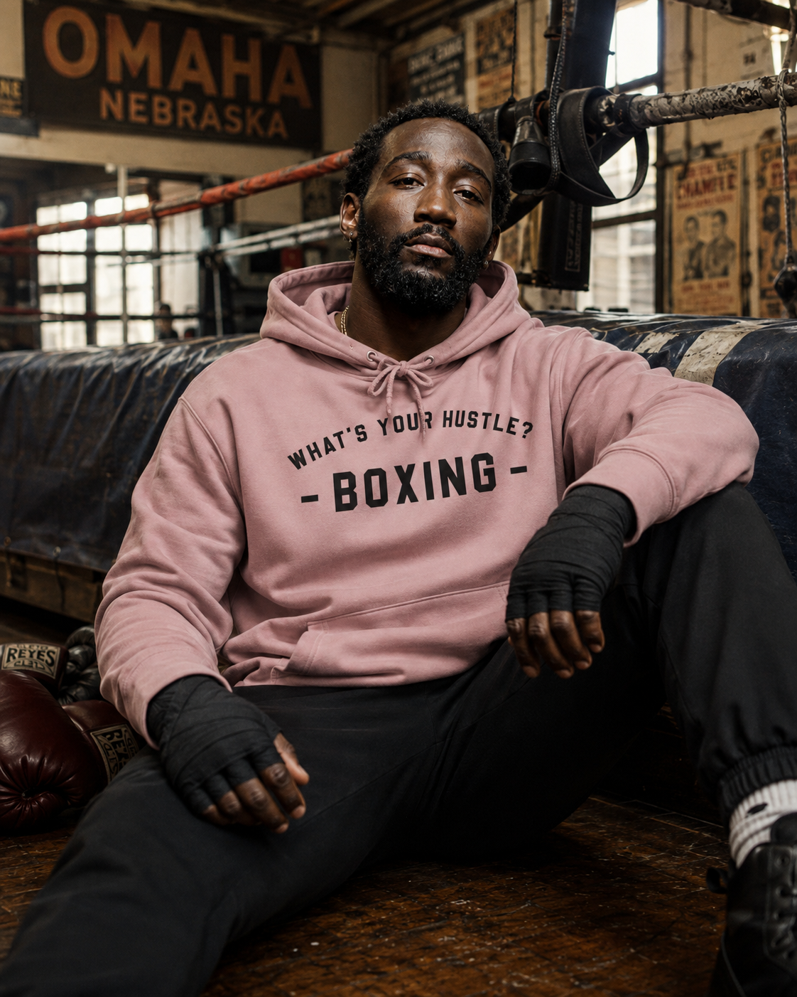A man sitting in a boxing gym wearing a dusty pink hoodie with 'WHAT'S YOUR HUSTLE? BOXING' printed in black across the chest, paired with black fingerless wraps and black sweatpants.
