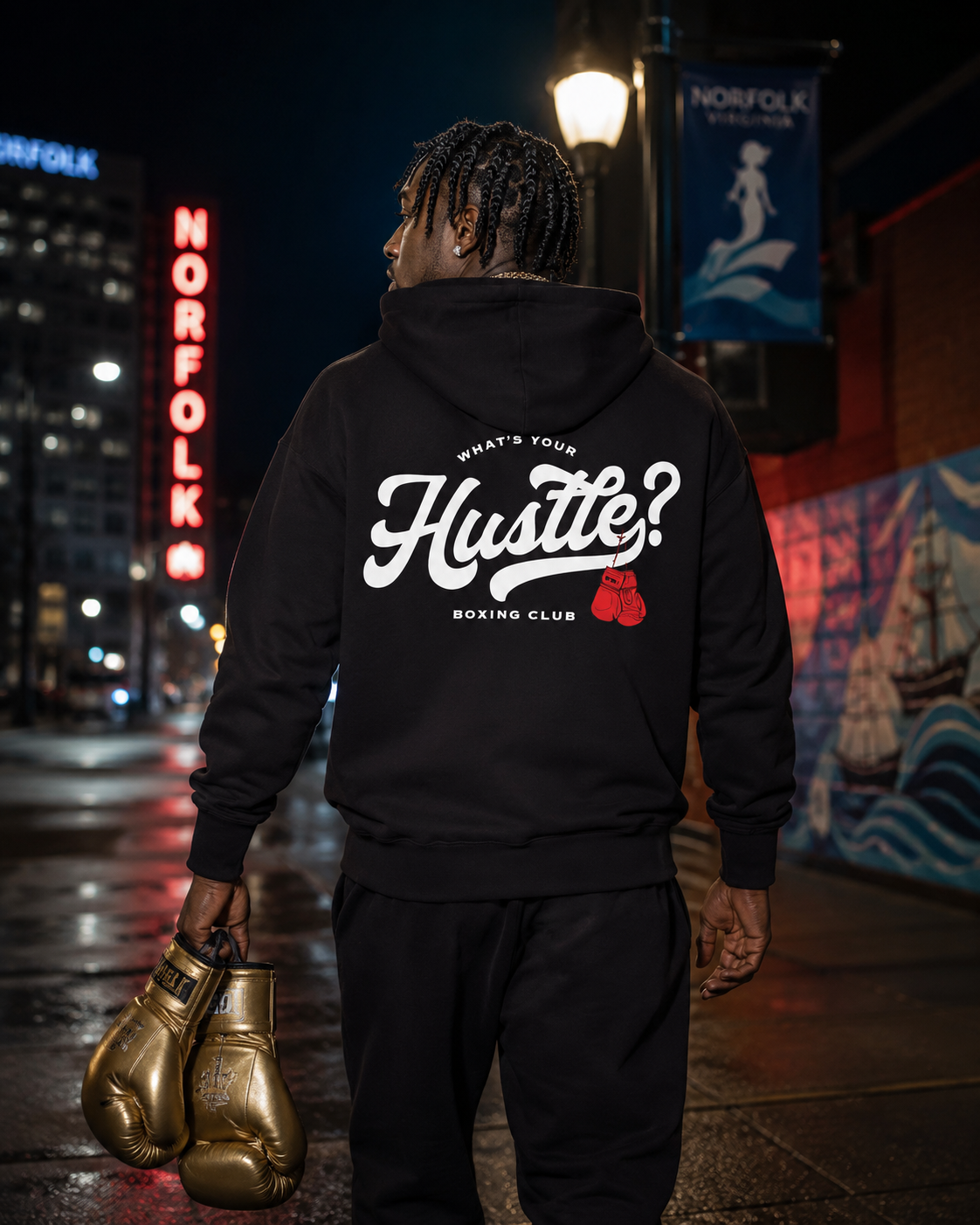A man stands on a city street at night wearing a black hoodie with a large white 'Hustle?' boxing graphic on the back, black sweatpants, and holding a pair of metallic gold boxing gloves. The background shows a blurred Norfolk city street scene with a lit sign and mural.